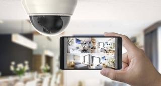 Une caméra IP fixée au plafond avec en arrière plan l&rsquo;intérieur de la demeure et au premier plan une main qui tient un smartphone sur lequel on peut voir les images prises par la caméra et qui correspondent à l&rsquo;intérieur du logement