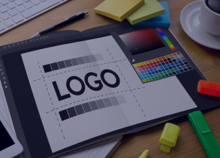 Bureau avec une ébauche de logo, un nuancier de couleur, des stabilo, trois bloc de post-it, un stylo et une tasse de café. On distingue un clavier de type Mac