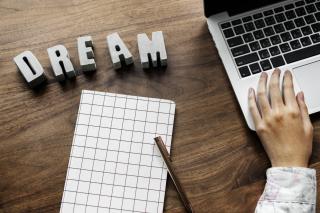 Bureau vue de dessus avec un carnet, une main d&rsquo;homme sur le clavier d&rsquo;un pc portable et le mot rêve en anglais: dream